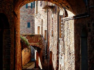 Standing in the cool shade under an archway inside a medieval walled town, looking through to bright sunshine highlighting the stone and brick walls of the buildings beyond. A second archway can be seen bridging two buildings