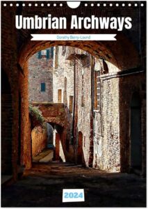 The front page of an A4 calendar showing a view underneath a dark archway towards a series of Italian traditional buildings bathed in sunlight. Has the words Umbrian Archways, Dorothy Berry-Lound and 2024.