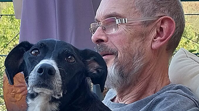 A smiling man with a beard and glasses sits with a black and white dog on his lap. The head of the man and the dog are visible and the dog is looking at the viewer.