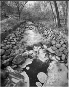 A black and white photo of a creek with sones along gthe edge and banks with trees. The surface of the water is covered with broken ice.