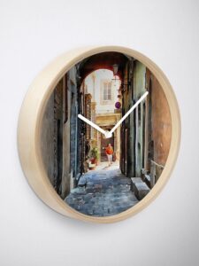 An angled view of a wall clock with bamboo frame and white clock hands. The image is of an alleyway in Cortona, Tuscany, with a man walking a dog.