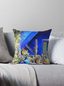 A square throw pillow sits on a grey sofa. The sofa is brigh and colorful, featuring blue a blue and yellow building and in front of it various cacti in coloured pots.