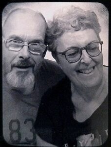 A black and white photograph with a vintage effect of a man and woman, both smiling and both wearing glasses